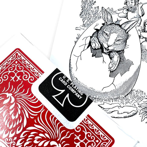 Kortos su raudonu ornamentiniu nugarėliu, matomas U.S. Playing Card Company lipdukas ir atidarytos dėžutės zuikučio iliustracija