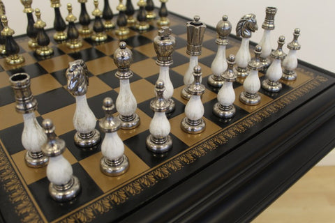 Šachmatų rinkinys: baltos medinės ir sendinto sidabro figūros ant juodo‑auksinės lentos, arti priekinio kampo