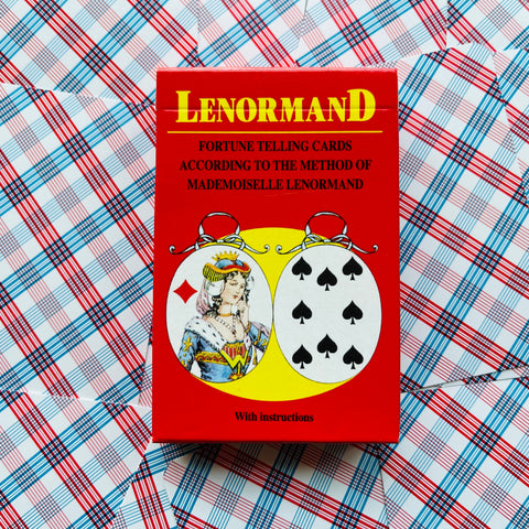 Lenormand kortų rinkinys raudoname dėkle su priekiu “With instructions”, plokščiai išdėstytas ant raštuoto languoto fono