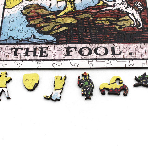 Dėlionė su mediniais teminiais gabalėliais, geltoni ir marginti mini figūrėlės bei „The Fool“ rėmo kraštas iš arti