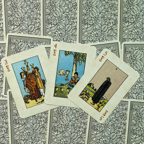 Tarot kortos Rider–Waite taurės: trys atvertos kortos (III, IV, V taurės) išdėstytos ant juodai baltų gėlių nugarėlių fono
