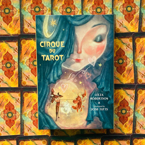 Cirque du Tarot kortų rinkinys su svajingu turkio–persikų viršeliu, moters profilio ir cirko figūrėlių iliustracija, padėtas ant kortų nugarėlių