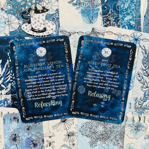 Arbatų kortelės, tamsiai mėlynos vaisių skonio etiketės dviem variantams (blueberry‑lemon ir berry), išdėstytos šone ant gėlių rašto fono