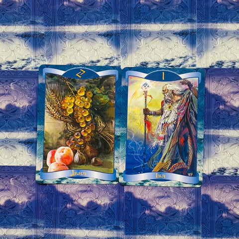Tarot kortos mėlyname rėme, dvi atverstos kortos (Jera ir Isa) ant ornamentuoto violetinio fono, saulės šešėliai