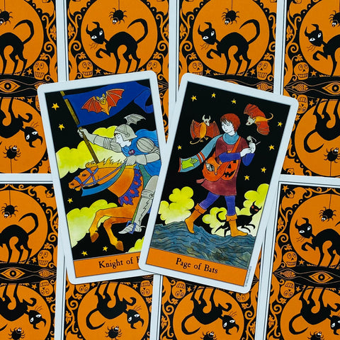 Tarot kortos su oranžinėmis nugarėlėmis ir juodu katės raštu — dvi atverstos kortos (Knight of Bats ir Page of Bats) matomos iš viršaus