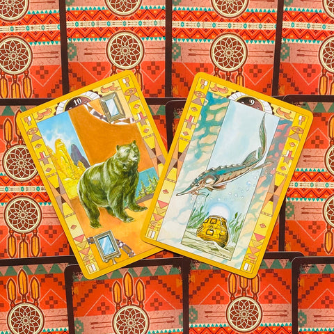 Tarot kortos su geltonais rėmeliais, dvi atverstos kortos (lokys ir ilgaūgė žuvis) ant raudono ornamentuoto fono