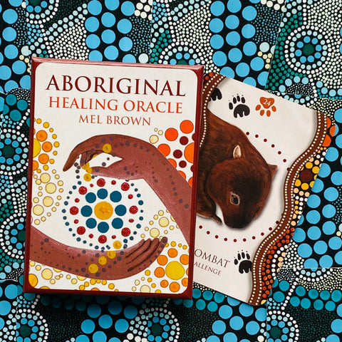 kortų rinkinys Aboriginal Healing Oracle (Mel Brown) su raudonu dėklu ir 24 kortomis, padėtas ant mėlyno taškuoto fono, pabrėžiantis dėžutės ir vienos kortos dizainą