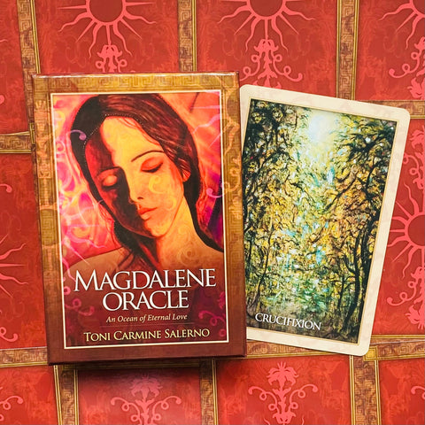 Magdalene Oracle kortos: raudona dėžutė su auksiniu rėmu ir šalia atversta korta „Crucifixion“ ant raudono ornamentuoto fono