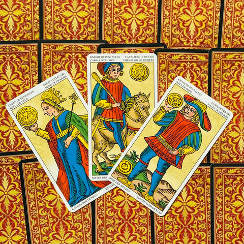 Tarot kortos Marseilio stiliaus — trys Pentaklių kortos (karalienė, riteris, jaunuolis) išsidėsčiusios ventyla ant raudono‑geltono raštuoto nugarėlių fono