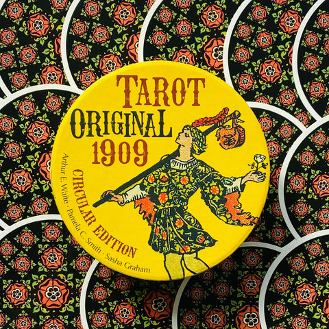 tarot kortos Original 1909 apvalioje geltonoje dėžutėje su klajūno iliustracija, matomi juodi gėlių rašto kortų nugarėlės