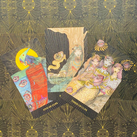 tarot kortos su rankų darbo iliustracijomis, atvertos kortos Penkių taurų ir Trijų taurų ant juodo‑auksuoto fono