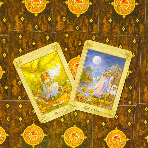 Tarot kortos — dvi atverstos kortos (Nine ir Eight of Cups) ant rudos‑auksinės nugarėlės, fotografuota iš viršaus, akcentuojamos iliustracijos