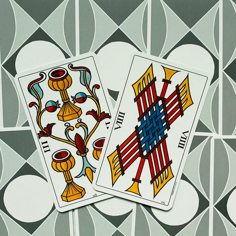 Tarot kortos didelio formato su ryškiomis geltonomis, raudonomis ir mėlynomis detalėmis, dvi pasvirusios kortos ant pilko geometrinio fono