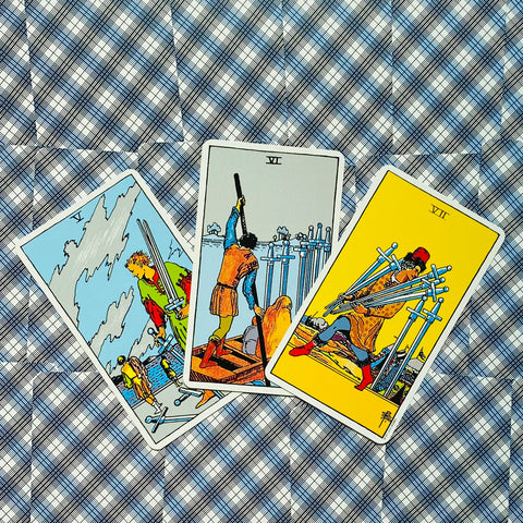 tarot kortos Rider–Waite trijų kardų variantas: ryškūs geltoni ir mėlyni piešiniai, išdėstyti kampu ant mėlyno langelėto audinio