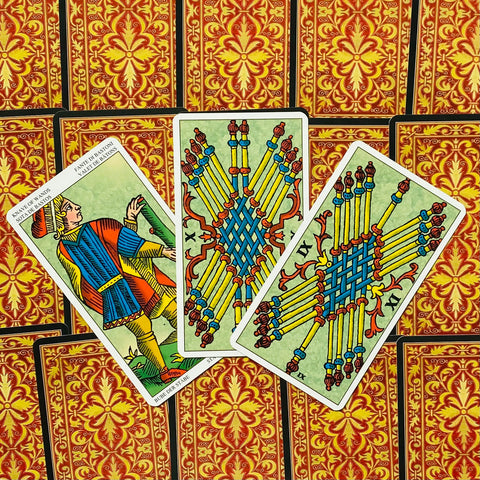 Tarot kortos Marseille stiliaus, ryškios geltonos–mėlynos lazdelės ant trijų atverstų kortų prie raudono‑auksinio nugarėlių rašto