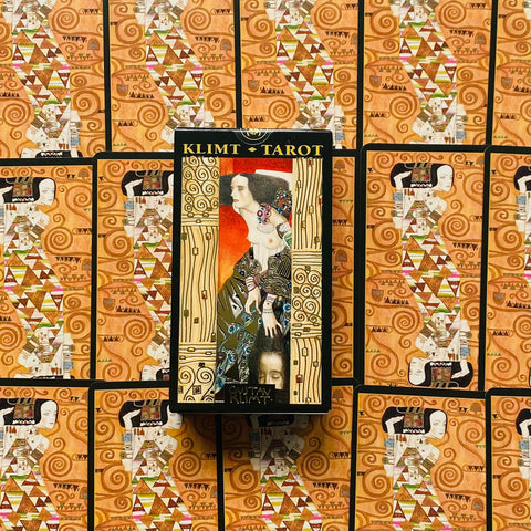 Klimto taro kortų rinkinys — dėžutės priekinė dalis ant derinančių kortų fono, pabrėžia auksinius ir oranžinius ornamentus