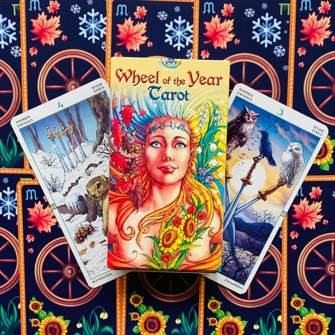 Tarot kortos Wheel of the Year – spalvinga dėžutė su moters iliustracija ir dviem kardų kortomis, fone ratai ir saulėgrąžos