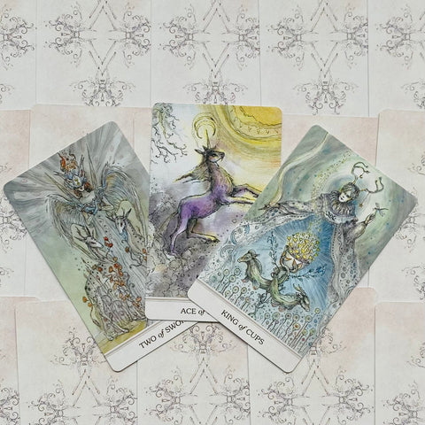 tarot kortos su akvarelinėmis iliustracijomis ir pastelinėmis spalvomis, trijų kortų atverstas fanas, matomas King of Cups ir Two of Swords