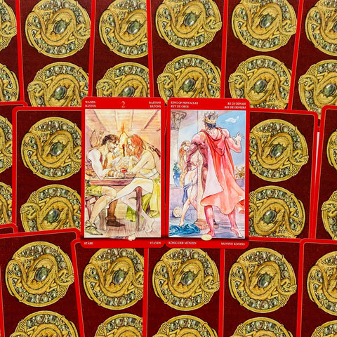 Tarot kortos su raudonomis nugarėlėmis ir auksiniu gyvatės ženklo raštu, dvi atverstos kortos centre ir artefaktinis maketas