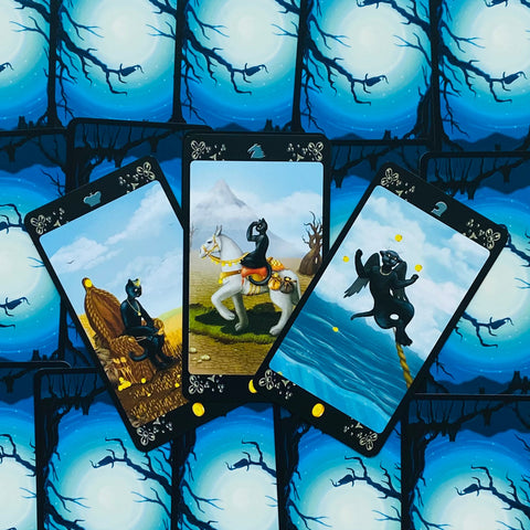 tarot kortos su juodos katės iliustracijomis — trys atverstos kortos (auksinės monetos, raitelis) ant mėlynos mėnulio nugarėlės