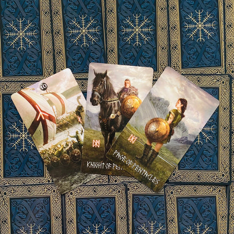 tarot kortos vikingų stiliaus rinkinys, trys atverstos kortos (Knight of Pentacles, Page of Pentacles) ant mėlyno ornamentuoto fono