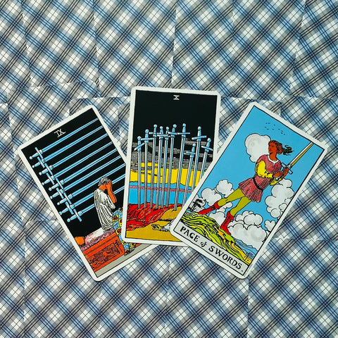 Tarot kortos trijų kortų išdėstymas — kardų devynetas, kardų dešimtukas ir Kardų Puslapis ant mėlyno languoto fono, ryškūs piešiniai ir kontrastingos spalvos