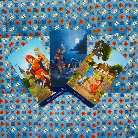 tarot kortos trijų kortų rinkinys: Six of Pentacles, Six of Swords ir Six of Cups ant mėlyno gėlėto fono, pakištos kampu