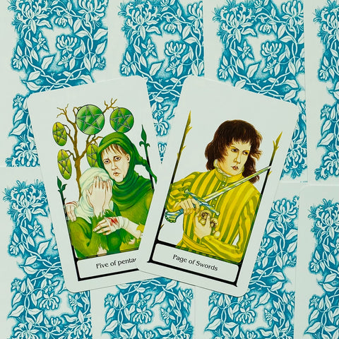 tarot kortos ant mėlyno gėlių dizaino fono, dvi kortos (Pentaklių penketukas ir Kardų puslapis) fotografuotos iš viršaus