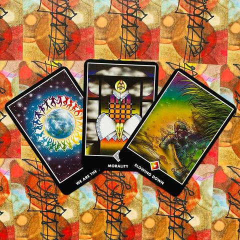 Tarot kortos trijų kortų išklotė: spalvingos „We Are The…“, Morality ir Slowing Down kortos ant raštuoto fono