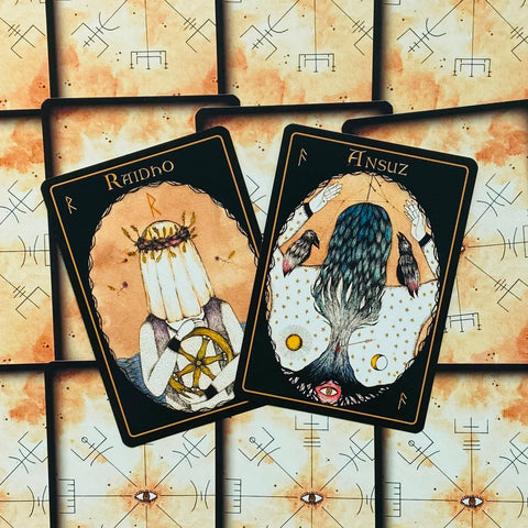 tarot kortos su juodais rėmeliais — artimas kampas, dvi atverstos iliustruotos kortos (Raidho, Ansuz) ant akvarelinių nugarėlių
