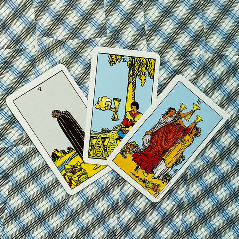 Tarot kortos trijų kortų išklotinė (taurų motyvai) ant mėlyno languoto audinio, kortos pasvirusios ir skirtingų variantų kompozicija