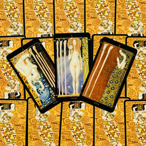 Tarot kortos su auksiniu ornamentu nugarėlėse, trys atvertos kortos išklotos centre ant raštuoto fono, rodoma kortų dizaino ir išdėstymo variacija
