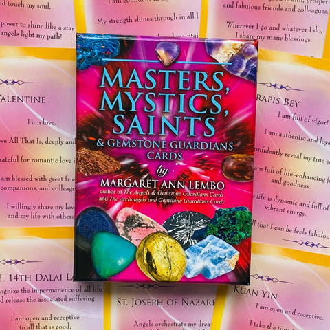 Kortų rinkinys Masters, Mystics, Saints ryškiai rožinėje dėžutėje su spalvotais akmenimis, padėtas ant išklotų afirmacinių kortelių