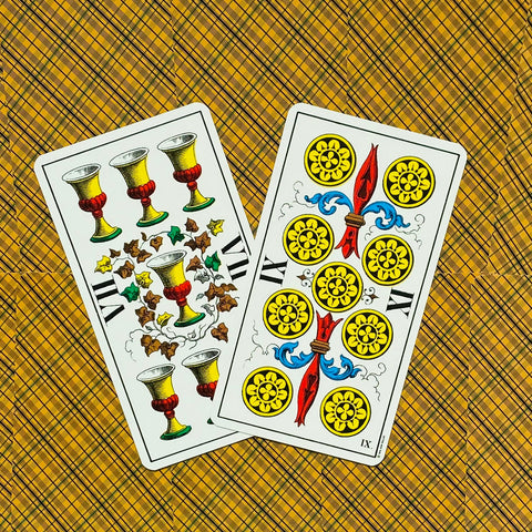Tarot kortos, dvi atverstos kortos (7 taurės ir 9 monetos) ant geltono raštuoto fono, matomi ryškūs piešiniai ir spalvos