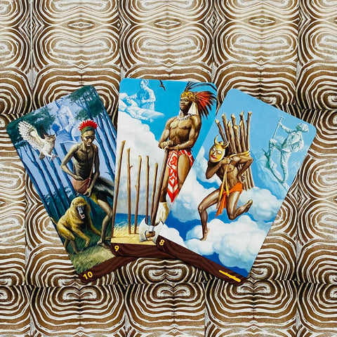tarot kortos su trimis spalvingomis iliustracijomis (8–10 lazgos), afrikietiški personažai ir debesų fonas, padėtos ant raštuoto rudo‑baltos audinio
