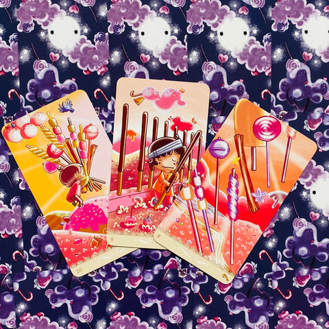 Tarot kortos su rausvai oranžinėmis saldainių tematikos iliustracijomis, trys kortos išskleistos ant tamsiai violetinio nugarėlių rašto, arti kadro ir skirtingų numerių variantai