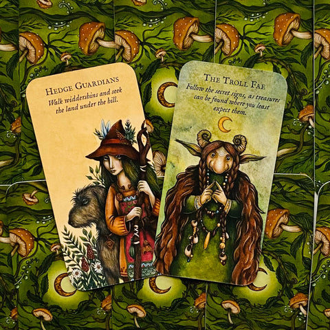 Kortos orakulo: dvi atverstos iliustruotos kortos (Hedge Guardians ir The Troll Fae) ant žalios grybiškos nugarėlės, arti kampo