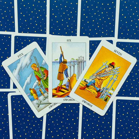 Tarot kortos Rider–Waite stiliaus, trys atverstos Kalavijų kortos (Penki, Šeši, Septyni) ant mėlyno žvaigždėto nugarėlio, centre fanuotos