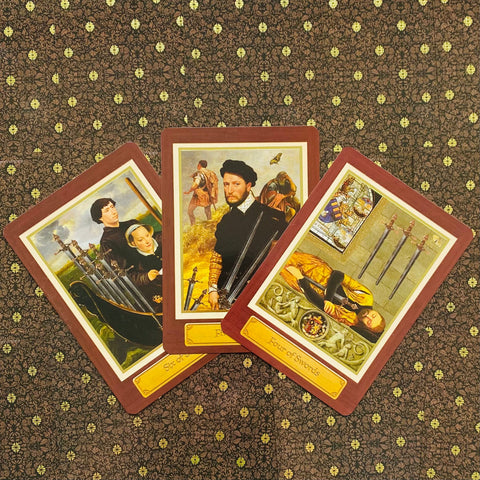 Tarot kortos rinkinys su rudu rėmeliu, trys atviros kortos išdėstytos vėduoklės forma ant raštuoto audinio fono