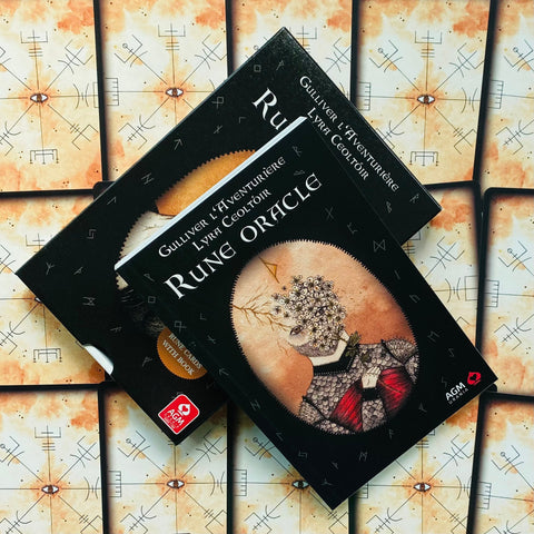 Rune Oracle rinkinys juodoje dėžutėje su knyga, AGM logotipu, rodomas kampu ant išdėstytų kortų nugarėlių