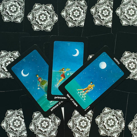 Tarot kortos mėlynomis iliustracijomis ir juodais kraštais, trys atviros kortos su mėnulio motyvais prieš ornamentuotą galinių kortų foną