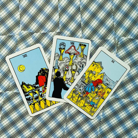 Tarot kortos Rider‑Waite rinkinys, trys taurės kortos išdėstytos vėduotai ant mėlyno‑baltos languotos medžiagos, matomas kortų priekis ir spalvotos detalės