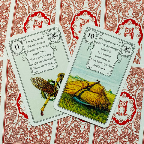 Lenormand kortos su raudonomis nugarėlėmis, dvi atvertos kortos – pjautuvas ir šluota, fotografuotos iš viršaus