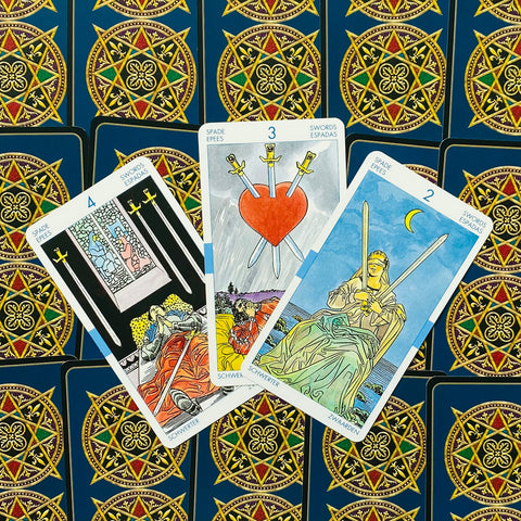 Tarot kortos Rider–Waite stiliaus, trys kardų kortos (2, 3, 4) išdėstytos vėduoklėje ant tamsiai mėlynos ornamentuotos nugarėlės