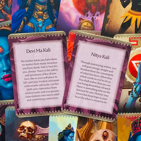 Orakulų kortos su violetinėmis rėmelėmis, artimas kadras atverstas į tekstą "Devi Ma Kali" ir "Nitya Kali", padėtos ant spalvingo iliustracinio fono