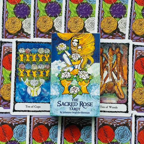 The Sacred Rose Tarot kortų dėžutė iš priekio su geltonomis taurėmis ir ilga moters šviesiaplauke, centre ant išdėstytų Ten of Cups ir Ten of Wands kortų, viršutinis žvelgiamasis kampas