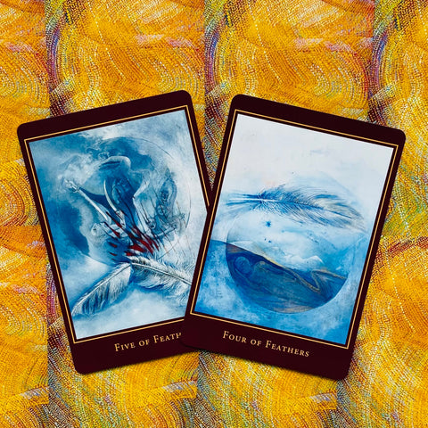 Tarot kortos su mėlynais Four of Feathers ir Five of Feathers iliustracijų variantais, persidengiančios ant geltono audinio