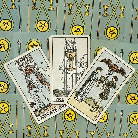 tarot kortos Rider–Waite stiliaus — trys atverstos kortos (Ace of Cups, King of Swords, Two of Cups) iš viršaus ant žalsvai melsvų nugarėlių rašto
