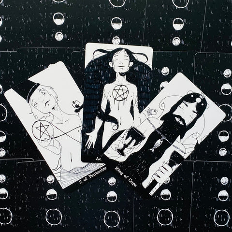 Tarot kortos juodai baltos iliustracinės, trys kortos (2 of Pentacles, Ace of Pentacles, King of Cups) išdėstytos vėduokle ant juodo rašto