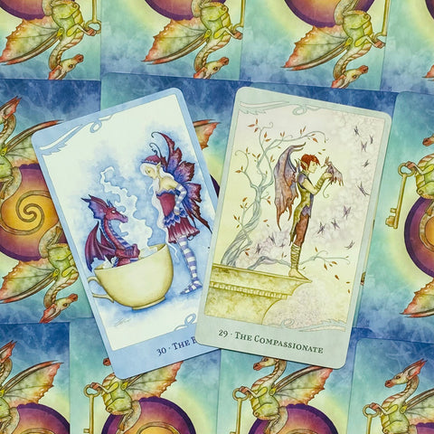 Tarot kortos su pastelinių mėlynų ir žalsvų atspalvių drakonų bei sparnuotų figūrų iliustracijomis, dvi skirtingos kortos parodytos iš priekio, skirtingos variacijos parduotuvės galerijoje